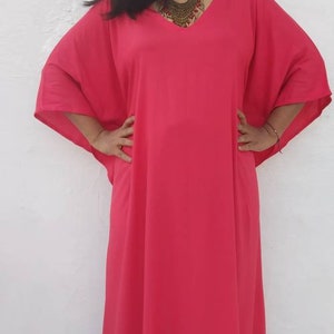 May include: A woman wearing a long, bright pink kaftan with a V-neckline and a gold necklace.