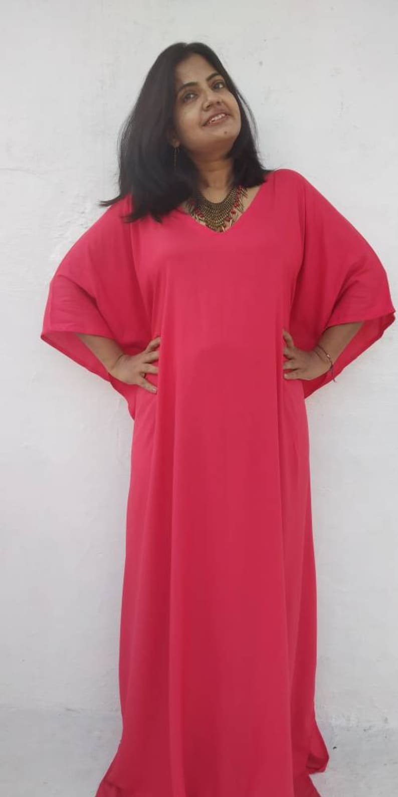 May include: A woman wearing a long, bright pink kaftan with a V-neckline and a gold necklace.
