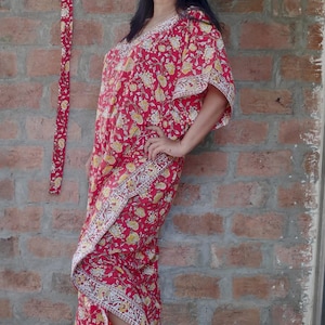 May include: A red and yellow floral print kaftan with a long, flowing skirt and a V-neckline. The kaftan is worn by a person standing in front of a brick wall.