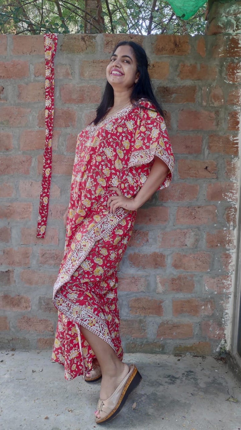 May include: A red and yellow floral print kaftan with a long, flowing skirt and a V-neckline. The kaftan is worn by a person standing in front of a brick wall.