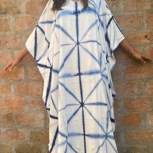 May include: A white caftan dress with a blue geometric tie-dye pattern. The dress has a V-neck and wide sleeves. The model is wearing pink sandals. The background is a brick wall.