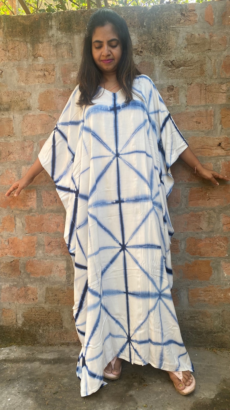 May include: A white caftan dress with a blue geometric tie-dye pattern. The dress has a V-neck and wide sleeves. The model is wearing pink sandals. The background is a brick wall.