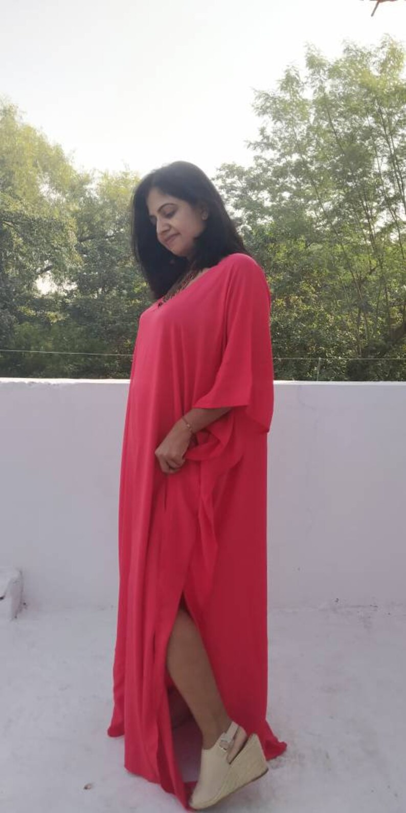 May include: A woman wearing a long, bright pink maxi dress with a side slit. The dress has a loose, flowing fit and is made of a lightweight fabric. She is wearing white wedge sandals.