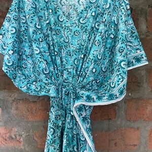 May include: A teal blue cotton kaftan with a white floral print. The kaftan has a V-neck and short sleeves. The fabric is soft and flowy.
