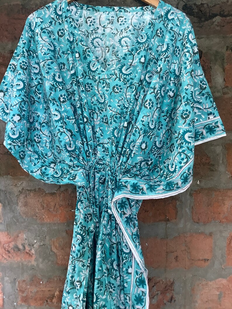 May include: A teal blue cotton kaftan with a white floral print. The kaftan has a V-neck and short sleeves. The fabric is soft and flowy.