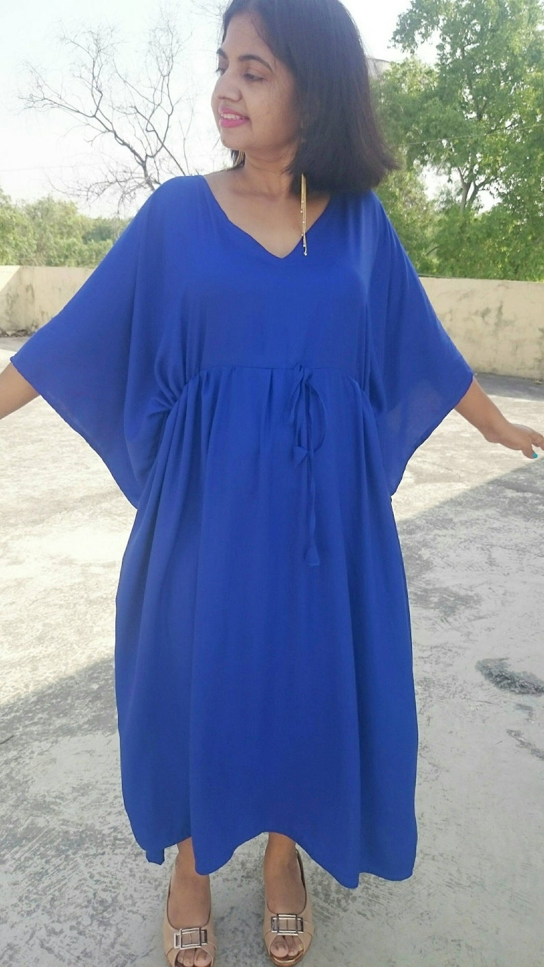May include: A vibrant blue caftan dress with a V-neckline and a drawstring waist. The dress has wide sleeves and falls to mid-calf. The person is wearing beige sandals. The background is a concrete surface with a tree in the distance.