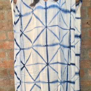 May include: A white caftan dress with a geometric blue tie-dye pattern. The dress has a V-neck and wide sleeves. The fabric appears lightweight and suitable for warm weather. The dress is floor-length.