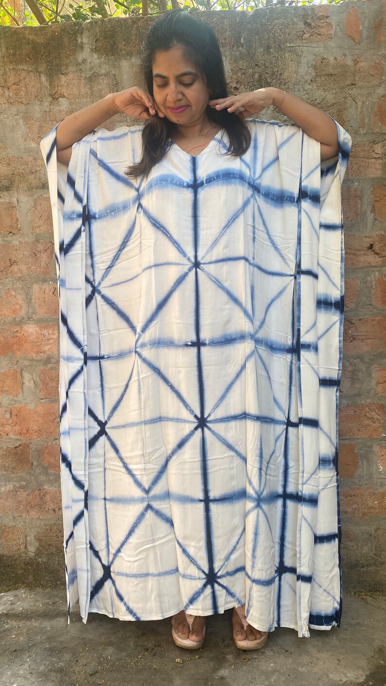 May include: A white caftan dress with a geometric blue tie-dye pattern. The dress has a V-neck and wide sleeves. The fabric appears lightweight and suitable for warm weather. The dress is floor-length.