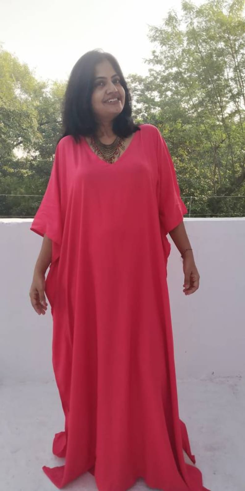 May include: A woman wearing a bright pink, long, loose-fitting kaftan with a V-neckline. She is wearing a gold necklace.
