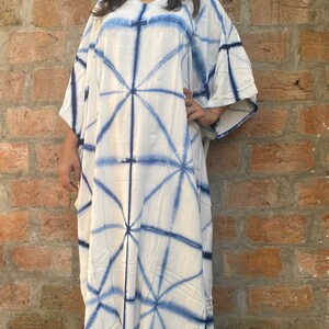May include: A white caftan dress with a blue geometric pattern. The dress has a V-neck and wide sleeves. The fabric appears to be lightweight, suitable for warm weather.