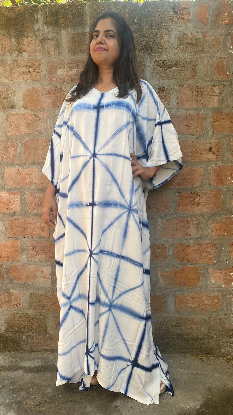 May include: A white caftan dress with a blue geometric pattern. The dress has a V-neck and wide sleeves. The fabric appears to be lightweight, suitable for warm weather.
