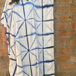 May include: White caftan dress with a geometric blue pattern. The dress has a loose, flowing design with wide sleeves. The blue pattern creates a grid-like design across the fabric. The dress is made of a lightweight material.