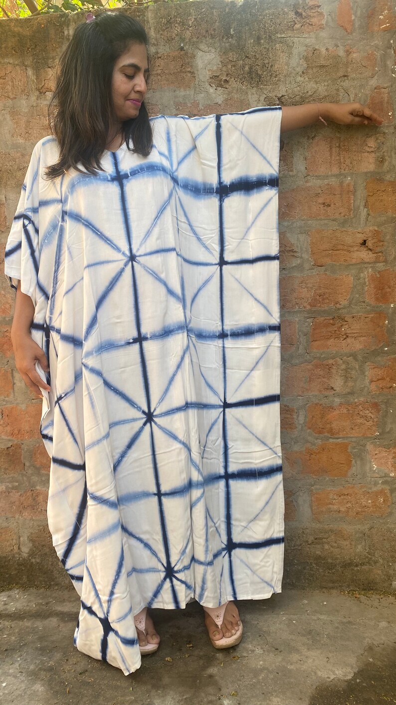May include: White caftan dress with a geometric blue pattern. The dress has a loose, flowing design with wide sleeves. The blue pattern creates a grid-like design across the fabric. The dress is made of a lightweight material.