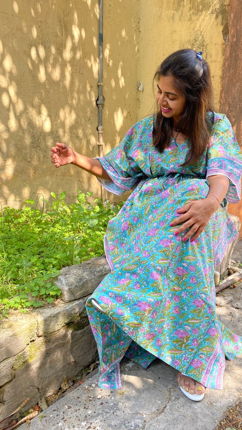 May include: A woman wearing a blue and pink floral print kaftan dress with white trim. The dress has a V-neck and long sleeves. She is sitting on a stone wall with green plants in the background.