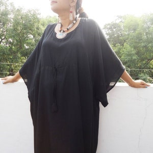 May include: A black maxi dress with a round neckline and long sleeves. The dress has a drawstring at the waist and a tassel detail. The model is wearing the dress with a silver and turquoise necklace and earrings.