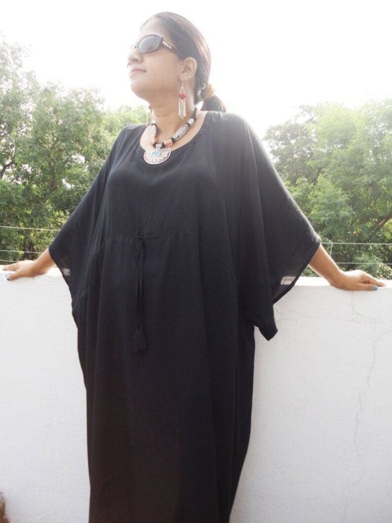 May include: A black maxi dress with a round neckline and long sleeves. The dress has a drawstring at the waist and a tassel detail. The model is wearing the dress with a silver and turquoise necklace and earrings.