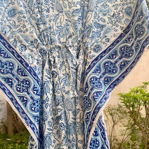 May include: A white kaftan dress featuring a blue floral and botanical print with a V-neckline and a drawstring waist. The garment includes wide sleeves and a decorative blue and white geometric border pattern along the edges of the fabric.