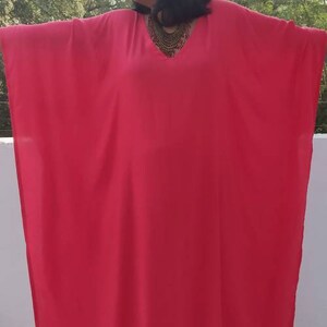 May include: A bright pink kaftan with a V-neckline and a gold necklace. The kaftan is made of a lightweight fabric and has a flowing silhouette.