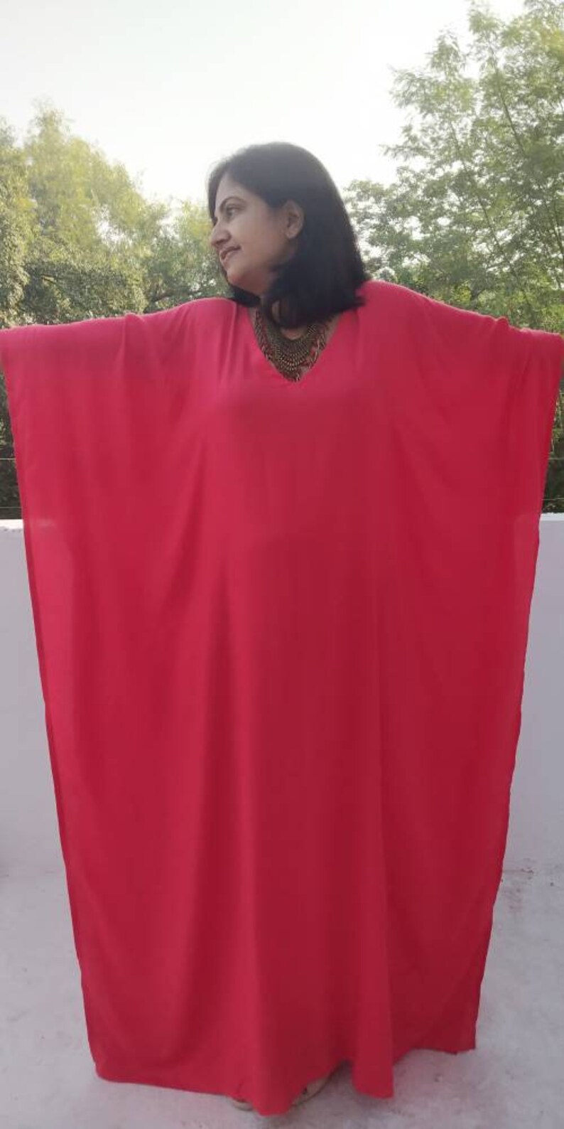 May include: A bright pink kaftan with a V-neckline and a gold necklace. The kaftan is made of a lightweight fabric and has a flowing silhouette.