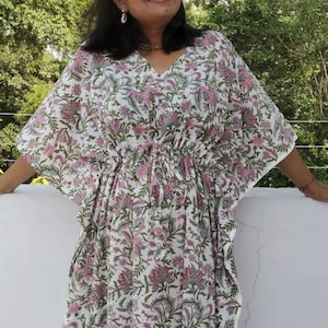 May include: A white cotton kaftan with a pink floral print. The kaftan has a V-neckline and a drawstring waist. The model is wearing the kaftan with a relaxed fit.