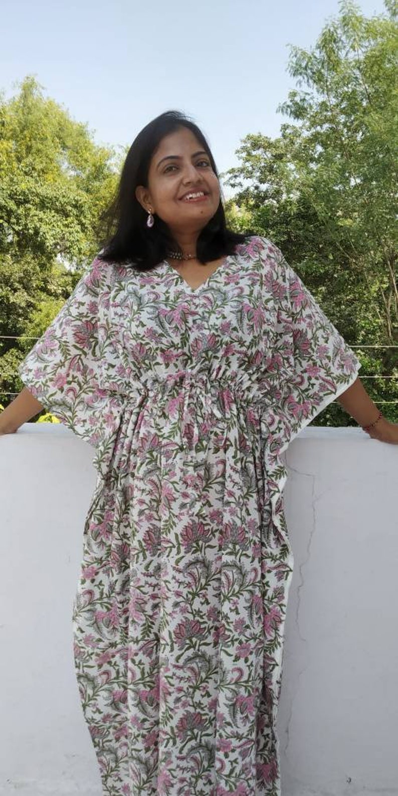 May include: A white cotton kaftan with a pink floral print. The kaftan has a V-neckline and a drawstring waist. The model is wearing the kaftan with a relaxed fit.