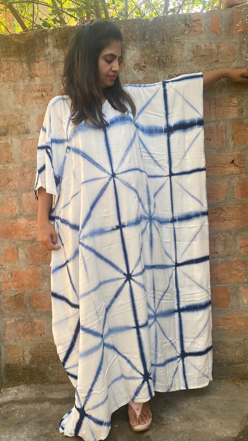 May include: A white, loose-fitting caftan dress featuring a blue geometric tie-dye design. The dress has wide sleeves and a relaxed silhouette. The fabric looks lightweight, ideal for warmer climates.