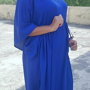 May include: A long, flowing cobalt blue dress with a V-neck and wide sleeves. The dress is cinched at the waist with a drawstring. The model is wearing gold earrings and sandals.
