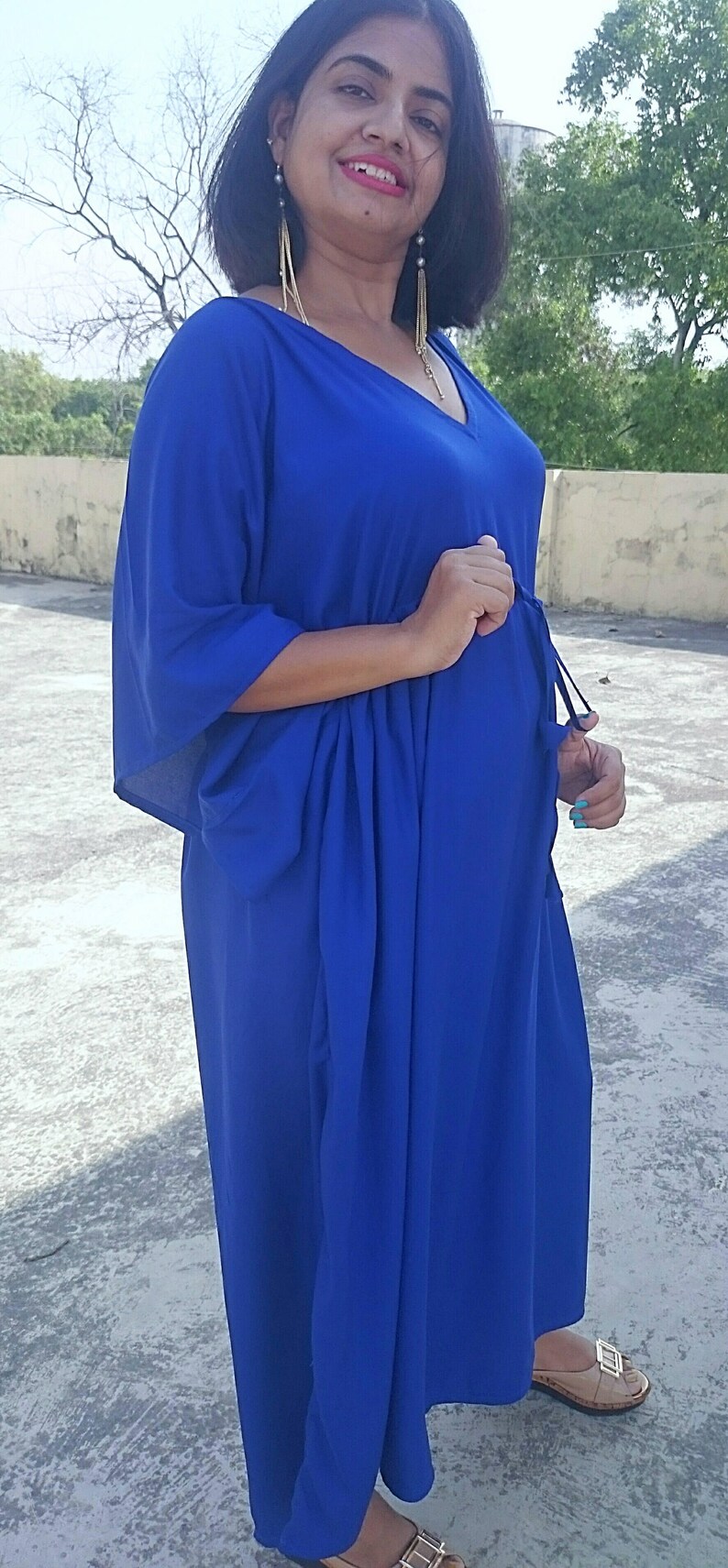 May include: A long, flowing cobalt blue dress with a V-neck and wide sleeves. The dress is cinched at the waist with a drawstring. The model is wearing gold earrings and sandals.