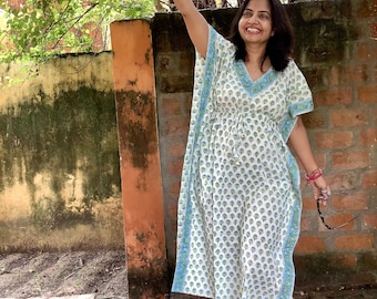 May include: A woman wearing a white and blue floral print kaftan with a blue trim. She is standing in front of a brick wall and has her arm raised.