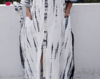 May include: A floor-length kaftan dress featuring a white base with vertical black tie-dye stripes. The garment has short, wide sleeves, a front button placket, and two large patch pockets on the front. The fabric has a loose, flowing silhouette.