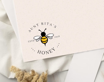Può includere: Un biglietto bianco con un logo di ape nero e giallo. Il logo dice "Aunt Rita's Honey" con "Estd 1946" sotto.