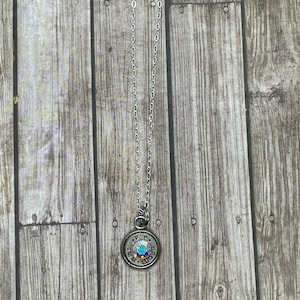 May include: A silver chain necklace with a round pendant featuring a 9mm Luger casing with a clear iridescent crystal in the center.