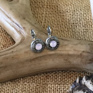 Peut inclure: Une paire de boucles d'oreilles en forme de balle argentées avec une pierre rose au centre. Les boucles d'oreilles sont gravées avec "9mm Luger" et "FC".