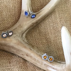 May include: A pair of silver bullet earrings with a blue, orange, and pink crystal. The earrings are displayed on a brown antler.