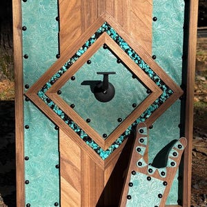 May include: A rectangular wooden wall display featuring a central diamond frame, turquoise stone accents, and teal embossed leather panels. The piece includes a black metal hook and a matching boot jack accessory, both secured with black rivets.