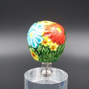 May include: A ceramic lamp finial with a floral design. The finial is painted with blue, yellow, red, and green flowers. It is sitting on a clear acrylic base.