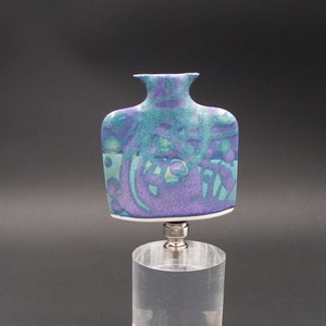 May include: A ceramic bottle stopper with a blue and purple abstract design. The stopper is on a clear acrylic base.