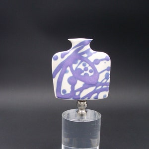 May include: White ceramic bottle stopper with a purple and blue abstract design. The stopper is on a clear acrylic base.