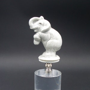 May include: A ceramic elephant figurine in a light gray color with a speckled finish. The figurine is standing on a round base and is attached to a metal lamp finial.