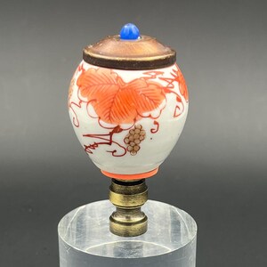 May include: A white ceramic lamp finial with a red and orange floral design and a blue top. The finial is on a clear acrylic base.