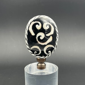 May include: Black and white glass finial with a swirling pattern. The finial is on a clear acrylic base.