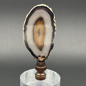 May include: A decorative lamp finial featuring a polished agate slice with a brown and cream center, mounted on a clear acrylic cylinder base. The agate has a dark outer edge and is attached to a bronze-colored metal fitting.