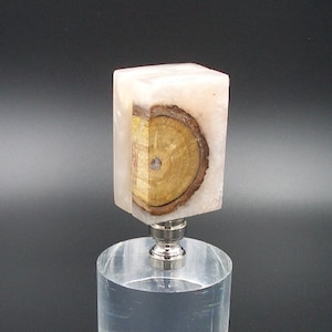 Puede incluir: Un remate de lámpara cuadrado hecho de resina rosa claro con una rodaja de madera incrustada en el centro. El remate está montado sobre una base de acrílico transparente.