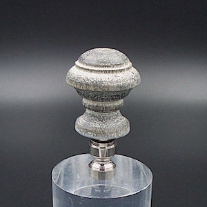 Puede incluir: Un remate de lámpara de madera gris con un diseño decorativo. El remate está montado sobre una base de acrílico transparente.