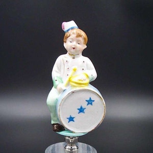 Puede incluir: Una figurita de porcelana de un niño con un uniforme blanco y verde y un sombrero rosa, sentado en un tambor azul y blanco con tres estrellas azules. La figurita está sobre una base de acrílico transparente.