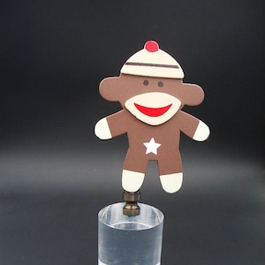 May include: A brown monkey lamp finial with a white and brown striped hat, red pom pom, white star, and a red smiling mouth. The finial is on a clear acrylic base.