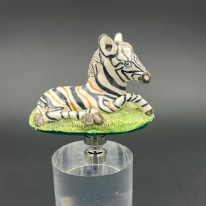 May include: A ceramic bottle stopper featuring a white and black zebra lying on a green grassy base. The zebra has orange stripes and blue eyes.