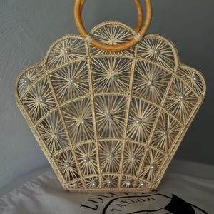 May include: A beige woven handbag with a fan-shaped design, decorated with small pearl accents. It has wooden handles and sits on a white cloth with a black logo. The bag's intricate pattern and natural tones create a stylish accessory.