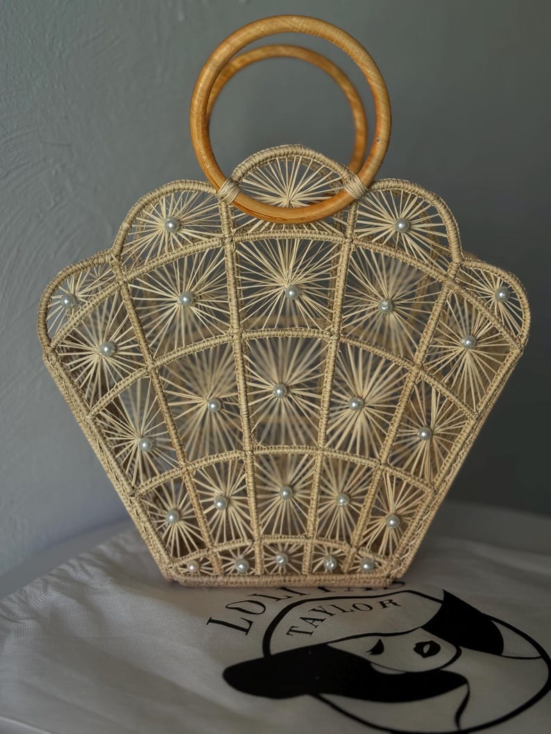 May include: A beige woven handbag with a fan-shaped design, decorated with small pearl accents. It has wooden handles and sits on a white cloth with a black logo. The bag's intricate pattern and natural tones create a stylish accessory.