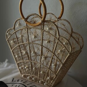 May include: A beige, woven handbag with a shell-like design, featuring wooden handles. The bag is adorned with small, white pearls and has a geometric pattern. The bag sits on a white surface.
